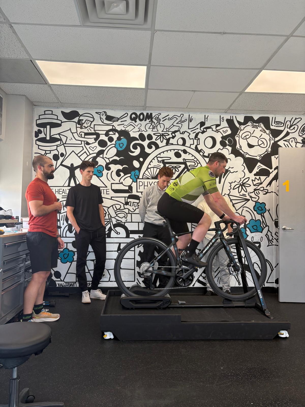 A rider pedalling on a trainer while the course group observes from the side